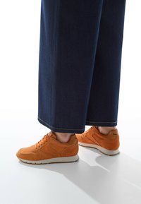 Baskets en daim orange avec des semelles blanches, présentant un design à lacets et des accents subtils. Associées à un pantalon en denim noir large.