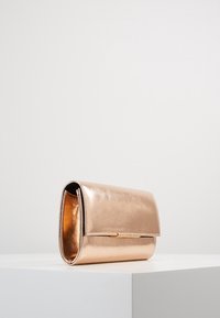 Bolso de mano metálico en oro rosa con cierre de solapa descansando sobre una superficie blanca contra un fondo gris claro.