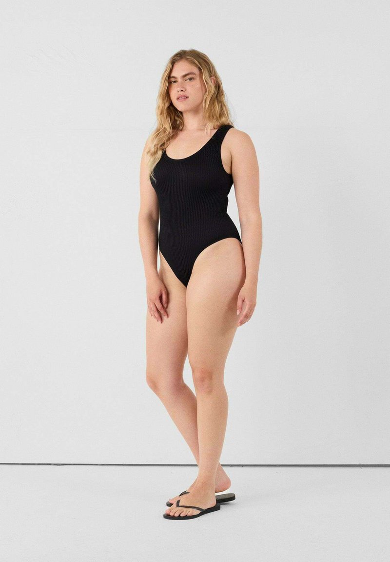 Zwart geribd badpak met een ronde halslijn, hoge beenopeningen en minimale bedekking. Gekoesterd met zwarte slippers.