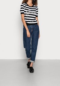 Czarnobiały w paski sweter z krótkim rękawem z napisem "Guess" w brokacie, zestawiony z niebieskimi dżinsami z podwiniętym dołem i czarnymi wsuwanymi butami.