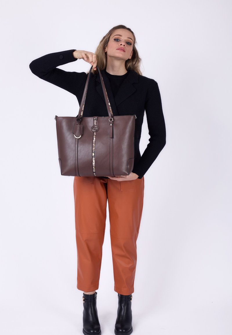 Kobieta w czarnym swetrze i brązowych spodniach trzyma dużą brązową skórzaną torebkę z wzorzystym pionowym paskiem i metalowym zawieszką.