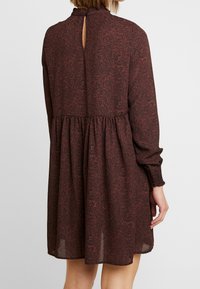 Kvinna som bär en lös, långärmad brun klänning med ett subtilt mörkt blommönster, insvängd midja och en liten nyckelhålsöppning vid bakre halsringningen.