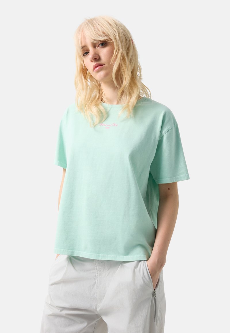 Blonde vrouw die een lichtmintgroen oversized T-shirt en losse witte broek draagt, met haar handen in de zakken tegen een effen achtergrond.