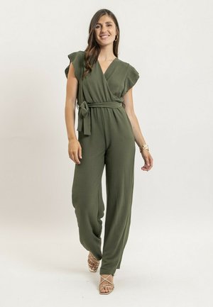 Femme souriante, portant une combinaison pantalon large de couleur olive avec des manches volantées et une ceinture à la taille, associée à des sandales à talons hauts à lanières.