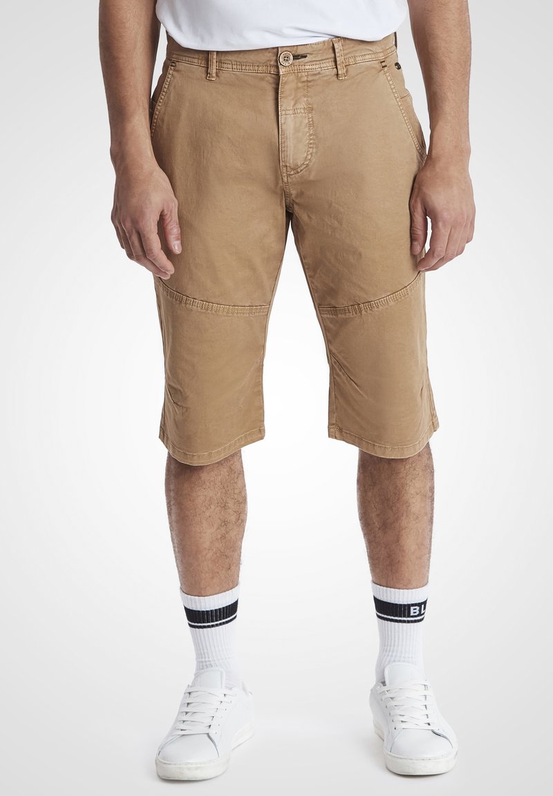 Beige shorts med avslappet passform, laget av bomull, kne-lengde, med kontraststing og sidelommer. Kombinert med hvite sneakers og stripete sokker.