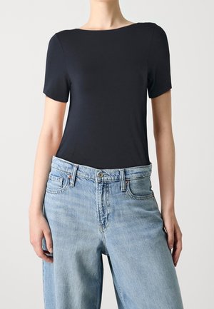 Osoba ubrana w dopasowaną czarną koszulkę z krótkim rękawem, wsuniętą w luźne, jasnoniebieskie dżinsy, stojąca na neutralnym tle.