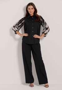 Schwarzes Hemd mit Knopfleiste und Blumen-Spitzenärmeln, kombiniert mit fließenden schwarzen Hosen und offenen schwarzen High Heels.