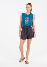 Top senza maniche color teal con stampa rosa, abbinato a pantaloni corti in lino grigio scuro con elastico in vita e cordoncino, indossato con infradito marroni.