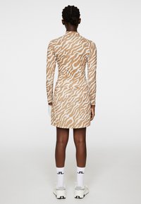 Långärmad, figurnära klänning i beige med vitt tigerrandigt mönster. Har hög krage och rynkdetalj vid midjan. Skorna inkluderar vita strumpor och sportskor.