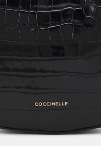 Czarna, błyszcząca torebka z teksturą krokodyla. Posiada złote logo "Coccinelle" w dolnej części. Gładka powierzchnia z wyraźnymi wzorami.