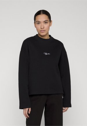 Kvinne med mørkt hår satt bakover, iført en svart ribbestrikket langermet topp og matchende bukser med liten hvit logotekst på brystet og beinet.