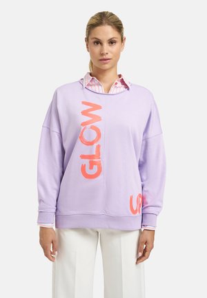 Woman wearing a light purple sweatshirt with "GLOW" in large orange letters and white pants, posing against a plain background.