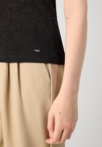 Black, textured top with metallic flecks, featuring a small, gold accent tag near the hem, paired with beige, pleated trousers.