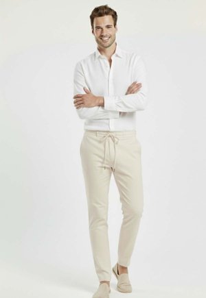 Hombre sonriente de pie con los brazos cruzados, vestido con una camisa blanca, pantalones beige con cordón y zapatos sin cordones a juego sobre un fondo blanco.