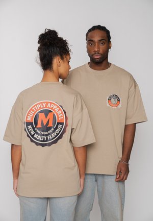 Mann und Frau tragen beige Multiply Apparel T-Shirts mit Logo auf der Vorderseite und großem kreisförmigem Design auf der Rückseite, stehen vor einem schlichten Hintergrund.