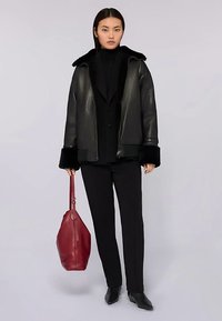 Černá kožená bunda se širokým límcem a kožíškovým lemem, doplněná černým topem a kalhotami. V ruce drží červenou koženou kabelku.