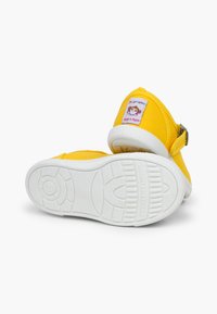 Zapatillas de lona amarillas con suela de goma blanca. Presentan un parche con el logo de un mono en el lateral y una correa con hebilla para un ajuste seguro.