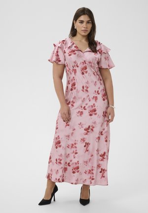 Femme portant une robe maxi fleurie rose avec des manches courtes à volants et des talons noirs à bout pointu, debout devant un fond clair uni.
