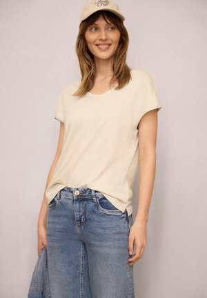 Smilende kvinde iført beige kasket, cremefarvet kortærmet top og blå jeans, holder en denimjakke i hånden mod ensfarvet baggrund.