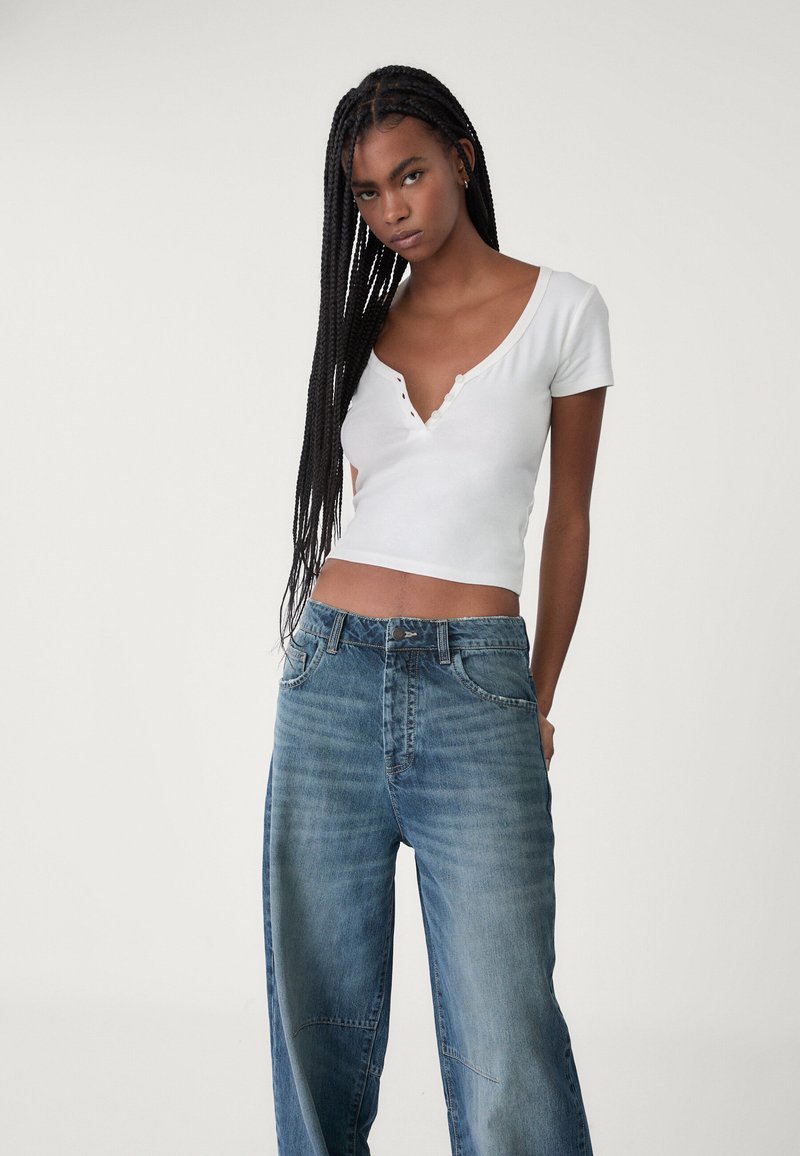 Vit kortärmad croptopp med djup V-ringning och knappdekorationer, kombinerad med högmidjade, ljusblå vida jeans.