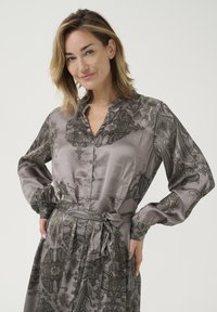Sukienka jedwabna w jasnoszarym kolorze z ciemnymi wzorami paisley, z zapięciem na guziki z przodu, długimi rękawami, falbaniastym kołnierzem i paskiem w talii do regulacji.