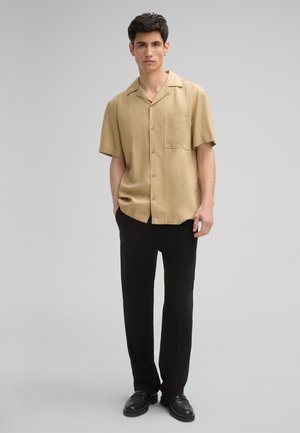Chemise beige à manches courtes avec une poche poitrine, accompagnée de pantalons noirs et de chaussures noires, sur un fond gris uni.