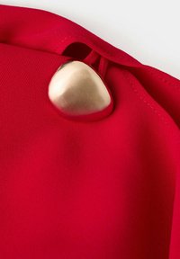 Fermeture à bouton métallique lisse sur tissu rouge vif avec coutures visibles et une petite boucle assurant le bouton.