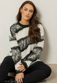 Frau mit langen braunen Haaren, die einen schwarz-weißen, gemusterten Pullover und schwarze Hosen trägt, sitzt im Schneidersitz gegen eine helle Wand.