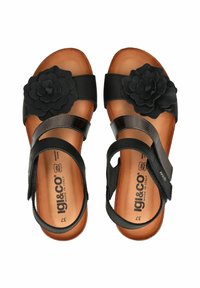 Black sandals featuring a large flower accent, two adjustable straps, and a cushioned insole with a tan sole. Made with leather material.