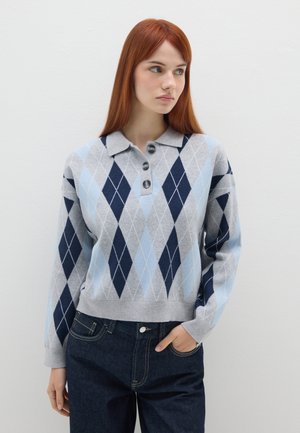 Pull en argyle gris, bleu et marine avec un col, trois boutons et un ourlet côtelé. Associé à un pantalon en denim foncé.