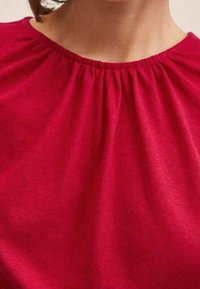 Rode blouse met een geplooide halslijn, gemaakt van zachte stof. De textuur is glad met een lichte glans. Geen zichtbare patronen of accenten.