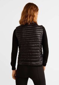 Schwarze gesteppte Weste mit horizontalen Nähten, leichtem Material, hohem Kragen und ohne sichtbare Verschlüsse, getragen über einem langärmligen schwarzen Oberteil.