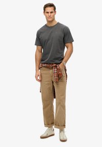 T-shirt grigio in cotone con scollo rotondo, pantaloni in corduroy color sabbia, camicia a quadri rossa e verde legata in vita, sneakers bianche con accenti beige.