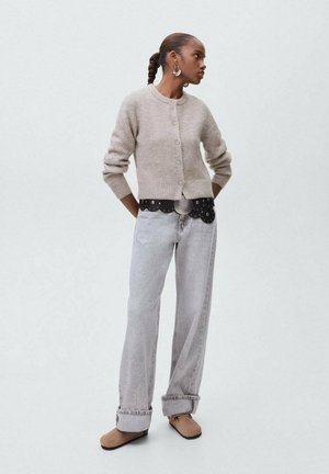 Femme avec des cheveux tressés portant un cardigan beige à boutons, une large ceinture noire, un jean gris clair avec des revers, et des chaussures glissantes marron, se tenant les mains derrière le dos.