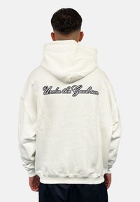 White hooded sweatshirt with a loose fit, featuring a textured "Under the Greek sun" embroidered detail on the back. Soft fabric, ribbed cuffs.