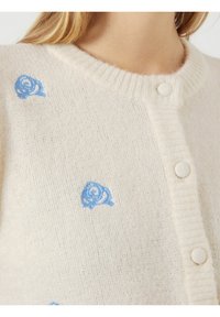 Kermakollane kardigan, mis on valmistatud pehmest kangast, millel on helesinised broderi roosid ja sobivad kaetud nööbid ees.