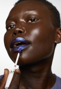 Rouge à lèvres liquide bleu vif appliqué avec un applicateur sur des lèvres brun foncé. Fini brillant avec une application lisse. Emballage transparent visible.