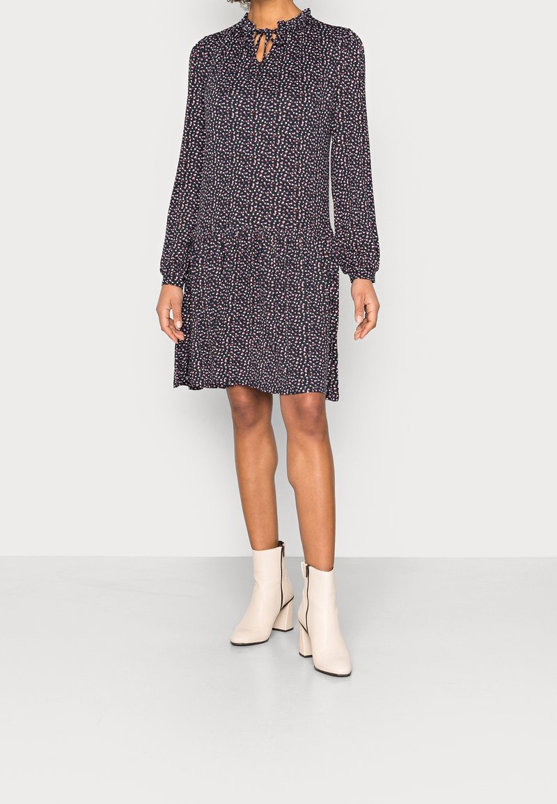 Femme portant une robe noire à manches longues avec des pois roses et blancs, un col à volants avec un lien, associée à des bottines écrues à talons blocs.