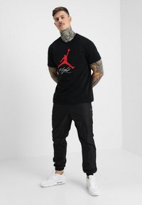 Svart T-skjorte med rødt "Jumpman"-logo og hvit "Flight"-tekst, kombinert med svarte joggebukser og hvite sneakers. Modellen har tatoveringer.