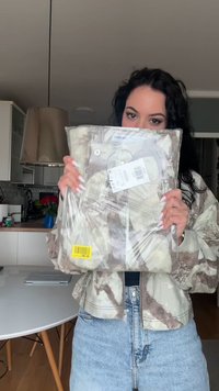 Žena s kovrčavom tamnom kosom drži novu jaknu u kamuflažnom uzorku u plastičnom pakiranju, stojeći u modernoj kuhinji s okruglim stolom.