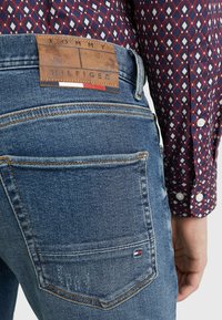 Blå denimjeans med en brun läderetikett med "TOMMY HILFIGER" präglat. Bakfickan har en liten flagg-emblem.