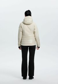 Puffet, lys beige jakke med hætte, har vandrette syninger, en blank finish og tætsiddende ærmer, parret med sorte bukser.