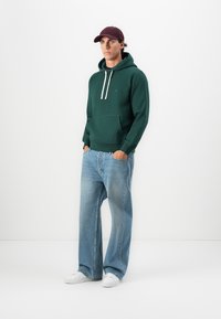Man wearing a dark green hoodie, light blue wide-leg jeans, white sneakers, and a dark red baseball cap, standing with hands in pockets.