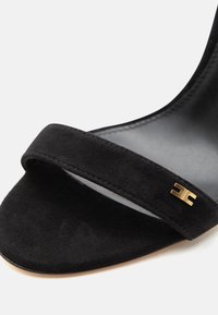 Μαύρο δερμάτινο σανδάλι με ανοιχτά δάχτυλα και έναν απλό ιμάντα με μικρή χρυσή διακοσμητική αγκράφα σε λευκό φόντο.