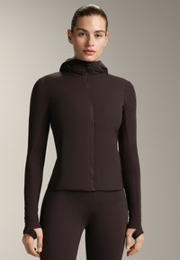 Brun zip-up jacka med en figursydd design och huva. Har långa ärmar med tumhål och är tillverkad av ett mjukt, stretchigt tyg.