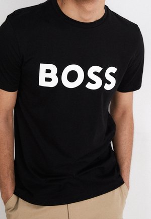Hombre con camiseta negra con el texto "BOSS" en blanco y en negrita, combinado con pantalones beige, con las manos en los bolsillos, frente a un fondo claro.