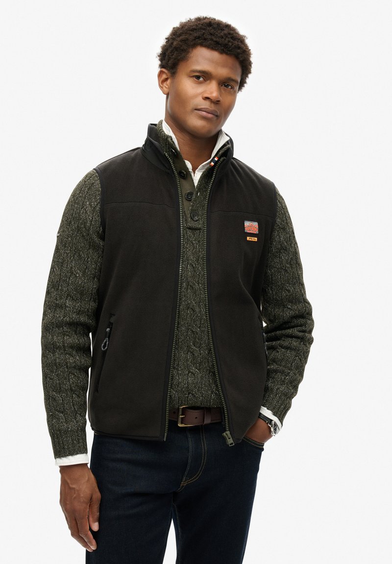 Gilet en polaire noir avec fermeture éclair à l'avant, poches latérales et logo. Porté par-dessus un pull en tricot torsadé vert, associé à un jean foncé et une ceinture marron.