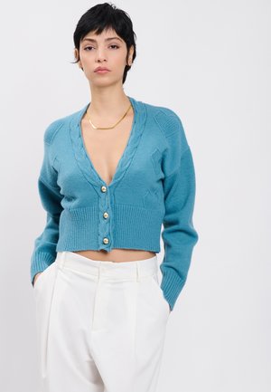 Frau mit kurzen dunklen Haaren, die eine blaue, bauchfreie Strickjacke mit Perlenknöpfen und weiße hoch taillierte Hosen trägt, steht vor einem schlichten Hintergrund.