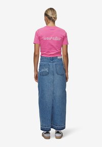 Rosa t-skjorte med korte ermer som har teksten "mest levende" på ryggen, parret med en blå denimmaxiskjørt og hvite sneakers.