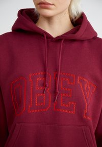 Κοντό μπουφάν μπορντό με μεγάλο κείμενο "OBEY" σε αντίθετη κόκκινη ραφή. Διαθέτει κουκούλα με κορδόνι και μπροστινή τσέπη τύπου καγκουρό.